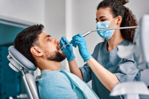 Male dental patient receiving treatment from a dentist wearing gloves and a mask, using dental instruments in a modern dental clinic.