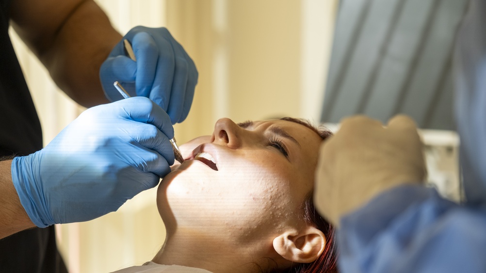 A skilled oral surgeon wearing surgical scrubs and a mask is carefully examining a patient's dental x-ray, with modern medical equipment visible in the background. No text on image.