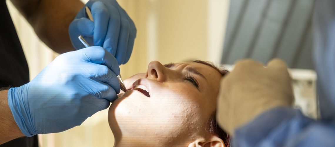 A skilled oral surgeon wearing surgical scrubs and a mask is carefully examining a patient's dental x-ray, with modern medical equipment visible in the background. No text on image.